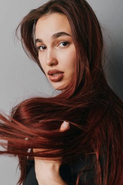 Düz kahverengi saçlı genç bir kadın kameraya bakarken saçlarına dokunuyor. Açık tenli güzel portre, ince makyaj ve tarafsız gri arka planda hafif ışıklandırma..