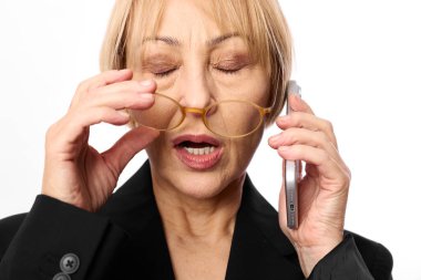 Siyah ceketli, kısa sarı saçlı orta yaşlı bir kadın gözlüğünü çıkarıp akıllı telefondan konuşuyor. Yorgun ve stresli görünüyor. Yorgunluk, iş stresi, iletişim ve çoklu görev anlayışı.