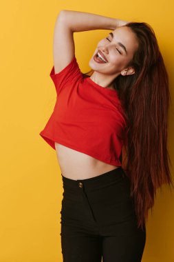 Kırmızı gömlekli, uzun kahverengi saçlı ve sarı arka planda poz veren siyah pantolonlu gülümseyen genç kadın. Kaygısız kadın modeli, mutluluk ve rahatlık gösteriyor. Gözleri kapalı ve elleri başının arkasında..