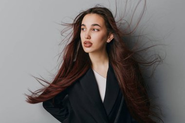Siyah ceket ve beyaz bluz giyen rüzgarlı saçlı genç bir kadının portresi. Tarafsız gri arkaplan, doğal görünüş ve yaşam tarzı ve güzellik temaları için zarif moda tarzıyla stüdyo çekimi.