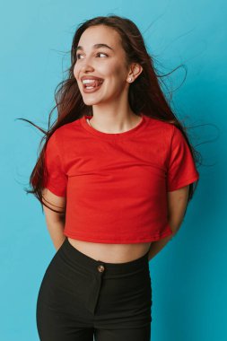 Kırmızı tişört ve mavi duvarda duran siyah pantolon giyen gülümseyen genç bir kadın. Uzun saç akışı, günlük kıyafet, neşeli ruh hali ve rahat yaşam tarzı ve moda için poz..
