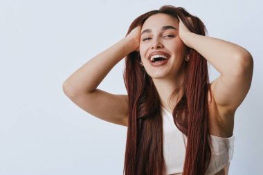 Uzun örgülü ve rahat duruşu olan gülümseyen kadın elleri kolsuz bluzuyla başını tutuyor. Pozitif duygu, gençlik ve tarafsız arka planda doğal ışık ile kaygısız tutum.