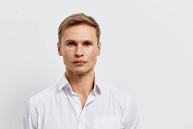 Açık renk saçlı, açık tenli, beyaz düğmeli gömlek giymiş sakin ve sakin bir ifadeyle kameraya bakan kendine güvenen bir gencin yakın portresi. Tarafsız bir arkaplanda stüdyo çekimi