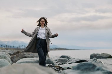 Beyaz gömlekli ve uzun paltolu bir kadın sakin suyu ve bulutlu gökyüzü olan kayalık sahil boyunca yürüyor. Gündelik sonbahar tarzı, doğa keşfi ve açık deniz manzarası teması.