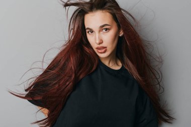 Siyah bluzlu, dalgalı kızıl saçlı genç bir kadının portresi gri duvarın önünde duruyor doğal güzelliğini ve kendine güvenini en az aksesuarlarla gösteriyor..