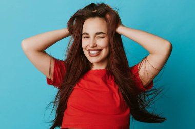 Uzun kahverengi saçlı, kırmızı gömlekli gülümseyen genç bir kadın mavi arka planda elleri saçlarında poz veriyor. Neşeli yüzü ve rahat duruşu olan bir model..