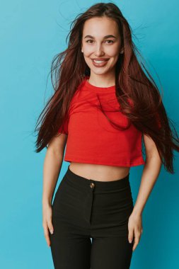 Kırmızı bluzlu gevşek kahverengi saçlı, gülümseyen kadın model ve mavi arka planda rahat poz veren siyah pantolon. Genç tarzı, mutluluğu ve kendine güveni olan modern kıyafetlerin içinde..