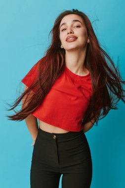 Kırmızı gömlekli ve mavi arka planda poz veren siyah pantolonlu kendinden emin genç bir kadın. Moda, güzellik, günlük tarz, uzun saçlı, gülümseyen ifade ve stüdyo resminde doğal neşeli ruh hali..