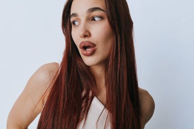 Kızıl saçlı, ağzı açık poz veren ve beyaz elbiseli genç bir kadın. Pürüzsüz ciltli kadın modelin yakın plan portresi ve açık arkaplanda gevşek bir ifade..