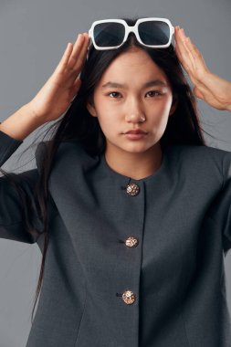 Saçı düz, beyaz güneş gözlüğünü stüdyoda kafasına takan şık bir kız. Gri ceketli ve kameraya tarafsız bir ifadeyle bakan genç bir kadın modelin portresi..