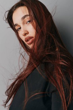 Siyah gömlek giyen, kırmızı saçlı ve az makyajlı genç bir kadının portresi. Sade gri bir arkaplanda doğrudan nötr bir ifadeyle bakarken saçları hafifçe hareket ediyor..