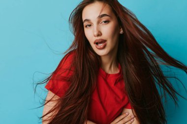 Uzun saçlı, kırmızı bluzlu çekici bir bayan model mavi arka planda poz veriyor. Yaşam tarzı, güzellik ve moda fotoğrafçılığı için saç hareketi, ifade ve günlük kıyafetler.