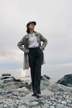 Gündelik kıyafetli genç bir kadın, elleri kalçasında çakıl taşı plajında poz veriyor. Bulutlu gökyüzü ve sakin su arka planı doğal ortamdaki gevşemiş ruh halini ve modern tarzı vurguluyor.