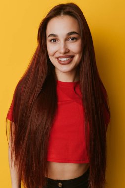 Uzun kahverengi saçlı, kırmızı bluzlu, sarı arka planda duran gülümseyen genç bir kadın. Parlak stüdyoda, doğal görünüşlü ve günlük kıyafetli neşeli bir kadının yakın plan portresi..