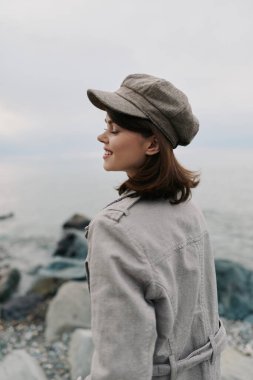 Sıcak ceket ve desenli şapka giyen gülümseyen kadın kayalık deniz kıyısında sakin bir anın tadını çıkarıyor. Yumuşak doğal ışık sahnenin sakinliğini ve rahatlığını arttırır..
