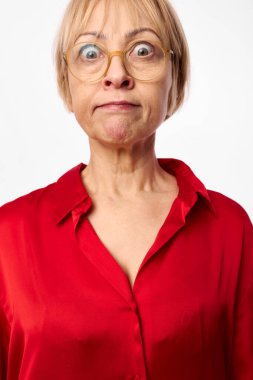 Gözlüklü, parlak kırmızı bluzlu, geniş gözlü ve büzüşmüş dudaklı yaşlı bir kadının portresi. Yüz ifadesi ve duygu konseptiyle beyaz arkaplan üzerine stüdyo çekimi.
