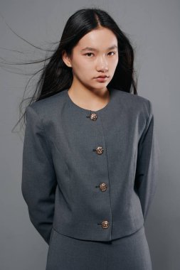 Düz siyah saçlı, gri ceketli, altın düğmeli genç bir kadının portresi. Tarafsız gri arkaplan ve moda konsepti için minimalist zarif tarzlı stüdyo fotoğrafı.