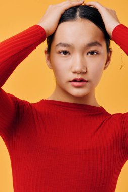 Kırmızı kazak giyen, elleri turuncu arka planda duran genç bir kadının portresi. Siyah saçlı Asyalı bir kadın model. İçerideki yumuşak ışık ayarlarıyla doğrudan kameraya bakıyor..