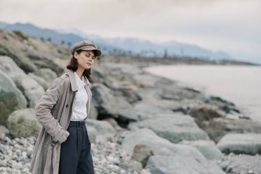 Gri paltolu ve şapkalı düşünceli genç bir kadın sakin bir su ve arka planda bulutlu bir gökyüzü olan kayalık bir kıyı şeridinde poz veriyor. Yalnızlık ve doğa kavramı.