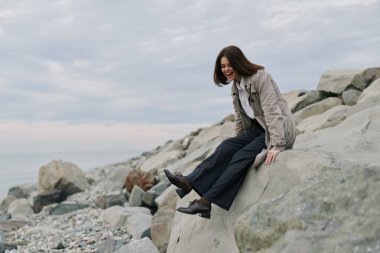 Kazaklı ve geniş pantolonlu mutlu bir kadın okyanus kıyısındaki kayalıklarda dinleniyor. Bulutlu gökyüzü ve doğal taşlar sakin, boş ve doğayla bağlantıyı vurguluyor.