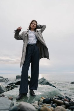 Ceket, gömlek ve geniş pantolon giyen şık bir kadın kayalık plajda rahatça poz veriyor. Bulutlu gökyüzü ve okyanus dalgaları doğal açık hava havası ve gündelik tarz kavramı ekler..