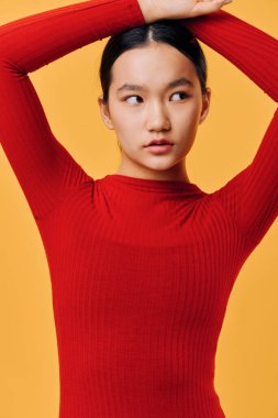 Kırmızı örgü süveter giyen zarif genç bir kadın. Kolları yukarıda, parlak sarı arka planda duruyor. Modern fotoğrafçılık için az makyajlı yumuşak tenli ve koyu renk saçlı bir kadın model..