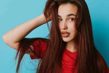 Uzun düz kahverengi saçlı, kırmızı bluzlu, elinde saçlarıyla mavi arka planda poz veren genç bir kadın portresi. Güzellik, moda, doğal görünüş ve kendine güvenen kadın..