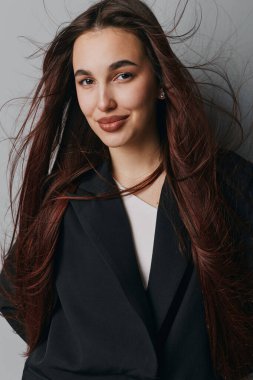 Siyah ceketli, beyaz tişörtlü, kendine güvenen genç bir kadın. Tarafsız gri arka planda rahat bir duruşla gülümsüyor. Sıradan stili ve hafif ışıklandırması olan güzel bir portre..