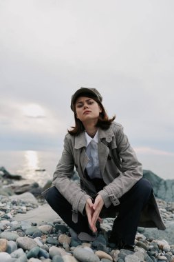 Kendinden emin genç bir kadın bulutlu gökyüzü ve sakin okyanusla kayalık kıyılarda paltosu ve şapkası ile poz veriyor. Sonbahar modası, açık hava yaşam tarzı ve doğal çevre kavramı.