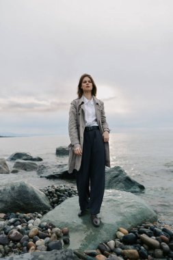 Uzun paltolu ve bol pantolonlu şık genç bir kadın Rocky Beach 'te duruyor. Bulutlu gökyüzü ve sakin su sakin bir atmosfer oluşturur. Tarz, doğa ve açık hava portresi kavramı.