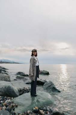 Paltolu ve şapkalı bir kadın bulutlu bir günde suyun kenarındaki kayanın üzerinde duruyor. Doğa kavramı, sakinlik, yalnızlık ve dış kıyı ortamında günlük moda..