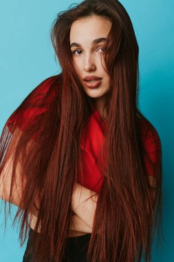 Uzun kahverengi saçlı, kırmızı bluzlu, kolları mavi arka planda çapraz duran genç bir kadın. Çekici bir kadın portresi. Stüdyoda doğal bir ifade ve rahat bir poz..