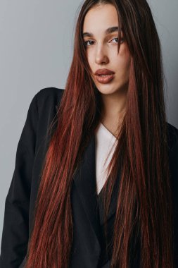 Siyah elbiseli, uzun kızıl saçlı kadın modelin portresi. Tarz ve zarafet için gri arka planda nötr bir ifadeyle kameraya bakıyor..