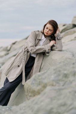 Bej ceketli kadın deniz kenarındaki kayalık araziye karşı duruyor. Bulutlu hava huzurlu atmosferi güçlendirir. Doğada moda, tefekkür ve mevsimsel stil kavramı.