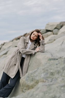 Şekli desenli bir palto giyen şık genç bir kadın bulutlu gökyüzünün altındaki kayalık araziye yaslanıyor. Huzurlu ruh hali stili ve doğa manzarasını ön plana çıkarıyor..