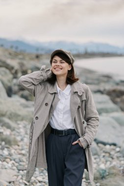 Paltolu ve şapkalı mutlu kadın kayalık sahilde elleri başının arkasında poz veriyor. Sonbahar modası, günlük giysiler ve doğal açık hava manzarası sakin ve hoş bir atmosfer yaratır..