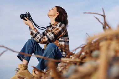 Sıradan giyinmiş bir kadın elinde bir kamera tutuyor, parlak gökyüzüne bakıyor, açık hava maceralarını ve doğa manzarasını kucaklıyor rahat bir hayat tarzı olan seyahat ve keşif anı.
