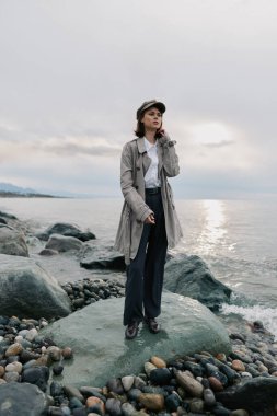 Uzun desenli palto ve şapka giyen şık bir kadın çakıl taşı plajındaki büyük kayanın üzerinde duruyor. Arka planda sakin bir deniz ve bulutlu bir gökyüzü var. Yalnızlık ve stil taşıyor..