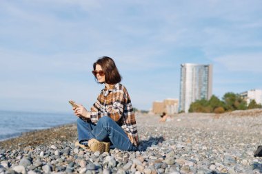Sahil kenarındaki çakıl taşlarının üzerine oturmuş ekose gömlek ve kot pantolon giymiş, akıllı bir telefonla göz gezdiren sakin bir sahil manzarası. Parlak gökyüzünün altındaki kentsel ufuk çizgisi..
