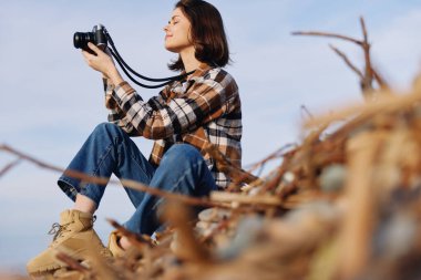 Çimlerin ve tarlaların arasına tünemiş bir kadın fotoğrafçının olduğu açık hava sahnesi, kamera hazır, sakin bir odak, ufuk ve yaratıcı çerçeveyle doğa ve manzaranın görüntüsünü bekliyor.