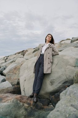 Trençkot giyen kadın kıyı şeridi yakınlarındaki kayalıklarda dikiliyor ve gökyüzü bulutlu. Doğal manzaralı açık hava sahnesi, sıradan moda, ve deniz kenarında barışçıl bir ifade..