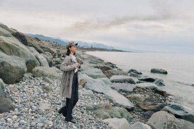 Paltolu ve şapkalı genç bir kadın kayalık kıyı şeridinde kamera tutuyor. Arka planda bulutlu hava ve sakin deniz açık hava fotoğrafçılığı ve doğa macerası temasını vurguluyor.