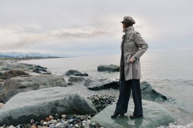 Gri paltolu ve şapkalı şık bir kadın kayalık sahilde denize bakıyor. Bulutlu gökyüzü ve nazik dalgalar sakin ve düşünceli bir ruh hali oluşturdu..