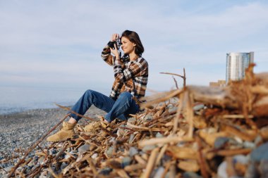 Kayalık bir plajda kadın fotoğrafçı bir kamera, sürüklenen bir ön plan, mavi gökyüzü, sakin bir sahne, doğa yolculuğu, gizli çekim, kıyı tarzı, çerçeve içinde hikaye anlatma
