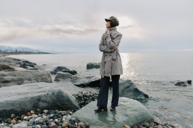 Sakin deniz ve kayalık sahil. Sıcak palto ve şapkalı kadın tek başına duruyor. Karamsar bulutlu gökyüzü ve uzak ufuklar yalnızlığı ve okyanusun yakınındaki düşünceli havayı vurgular..