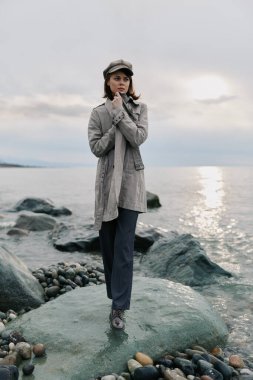 Sakin deniz kenarındaki kıyı kayalıklarında uzun gri palto ve şapka giyen bir kadın. Bulutlu gökyüzü ve yansıtıcı su dengesiz bir atmosfer yaratır. Açık hava tarzı ve mevsimlik kıyafet kavramı.