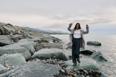 Okyanus dalgalarının yanındaki kayalık sahilde ceket ve pantolon giymiş biri. Sakin atmosfer taşları, suyu ve bulutlu gökyüzünü içeriyor, açık hava macerası ve doğal çevreyi vurguluyor..