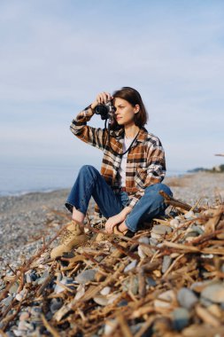 Bir çakıl taşlı plajda oturan fotoğrafçı, kamera yukarıda, kot pantolon ve ekose gömlek giyiyor, doğa, deniz esintisi ve gizli bir kıyı fotoğrafı sahnesine odaklanıyor..