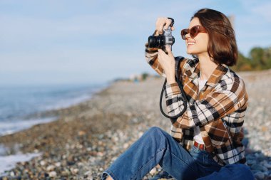 Güneşli bir sahilde kameralı bir kadının açık hava fotoğrafı, günlük kıyafetler, manzara ve portre çekimlerine odaklanmış, deniz kenarındaki anıları yakalıyor..