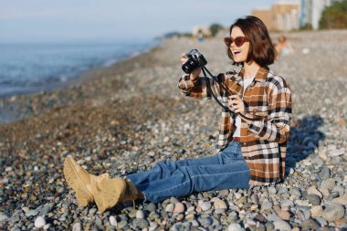 Çakıl taşlı bir plajda, elinde kamera, güneş gözlüğü ve ekose gömlek olan bir kadın fotoğrafçıyla açık hava çekimi, doğal ışık ve kıyı havasında gizli deniz resmi çekmek.